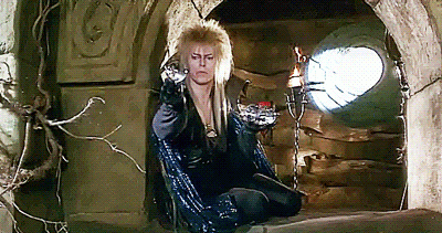 David Bowie dressed in a black cape and sparkling fantasy costume with a blonde spiky haircut, sitting in a castle window spinning crystal balls before releasing them onto the wind.