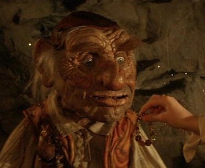 A wrinkly-faced dwarf man with a red leather skull-cap, orange waistcoast and white smock shirt. He looks amazed as a girl hands him a plastic bracelet.