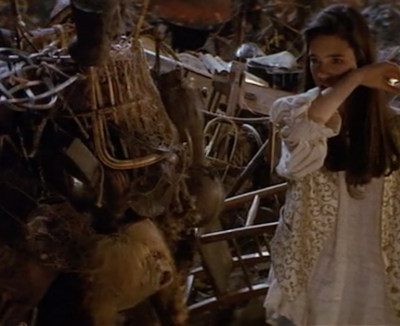 A teenage girl in a cream shirt and waistcoat stands in a junkyard. She is talking to a wrinkled old woman laden with junk.